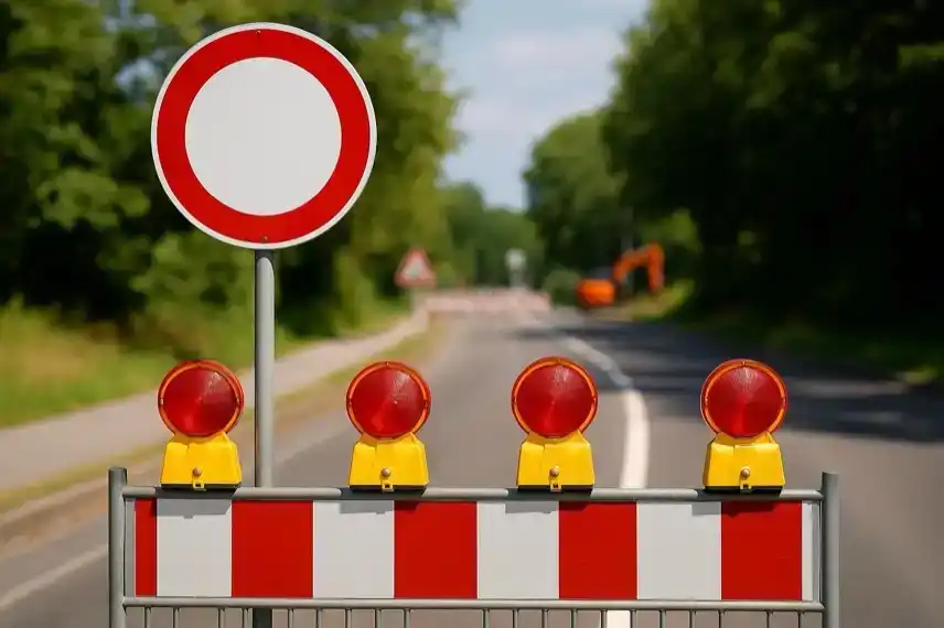 Baustellenabsperrung auf einer Landstraße mit roten Warnlichtern und Verkehrsschild im Vordergrund, Bäume und Straße im Hintergrund.