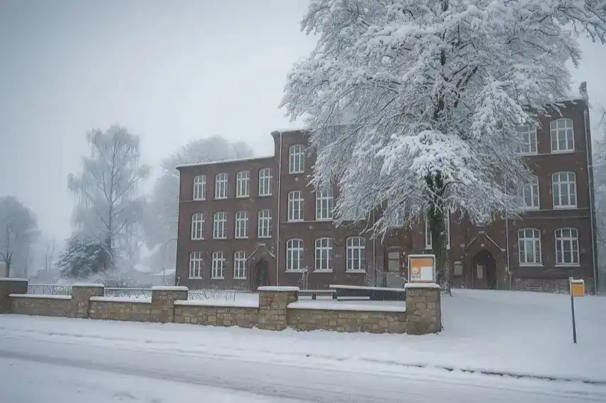 Rotes Backsteinschulgebäude mit verschneiten Bäumen und Steinmauer an einer ruhigen Straße im Winter.