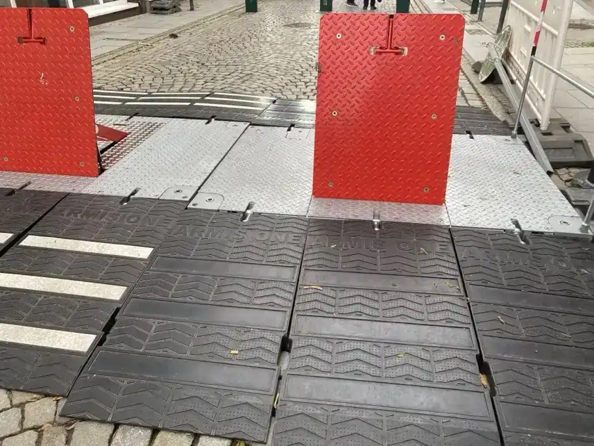 Poller mit rot-weißem Absperrband in einer historischen Altstadtstraße mit unscharfen Passanten im Hintergrund.