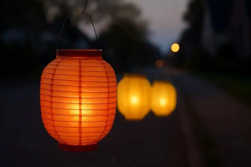 Drei bunte Papierlaternen leuchten in der Dämmerung auf einer Straße, im Hintergrund sind unscharf Häuser und Lichter zu sehen.