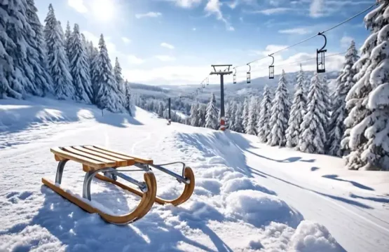 Verschneiter Hang mit Holzschlitten im Vordergrund, leere Sessellift-Anlage und schneebedeckte Bäume unter blauem Himmel bei Sonnenlicht.