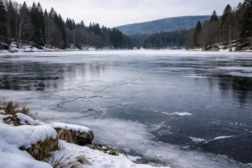 Zugefrorener See im Harz mit dünner Eisdecke, feinen Rissen und Schneerand am Ufer bei winterlicher Landschaft