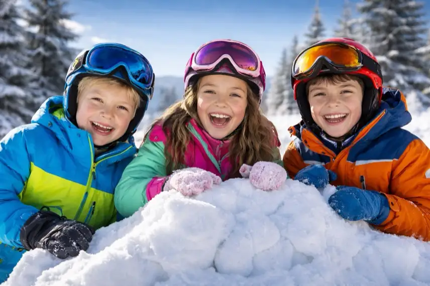 Drei lachende Kinder in bunten Skianzügen stehen vor einem selbst gebauten Schneeberg unter blauem Himmel.